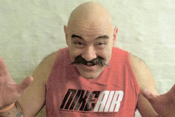 A bald man with a mustache is making a gesture.